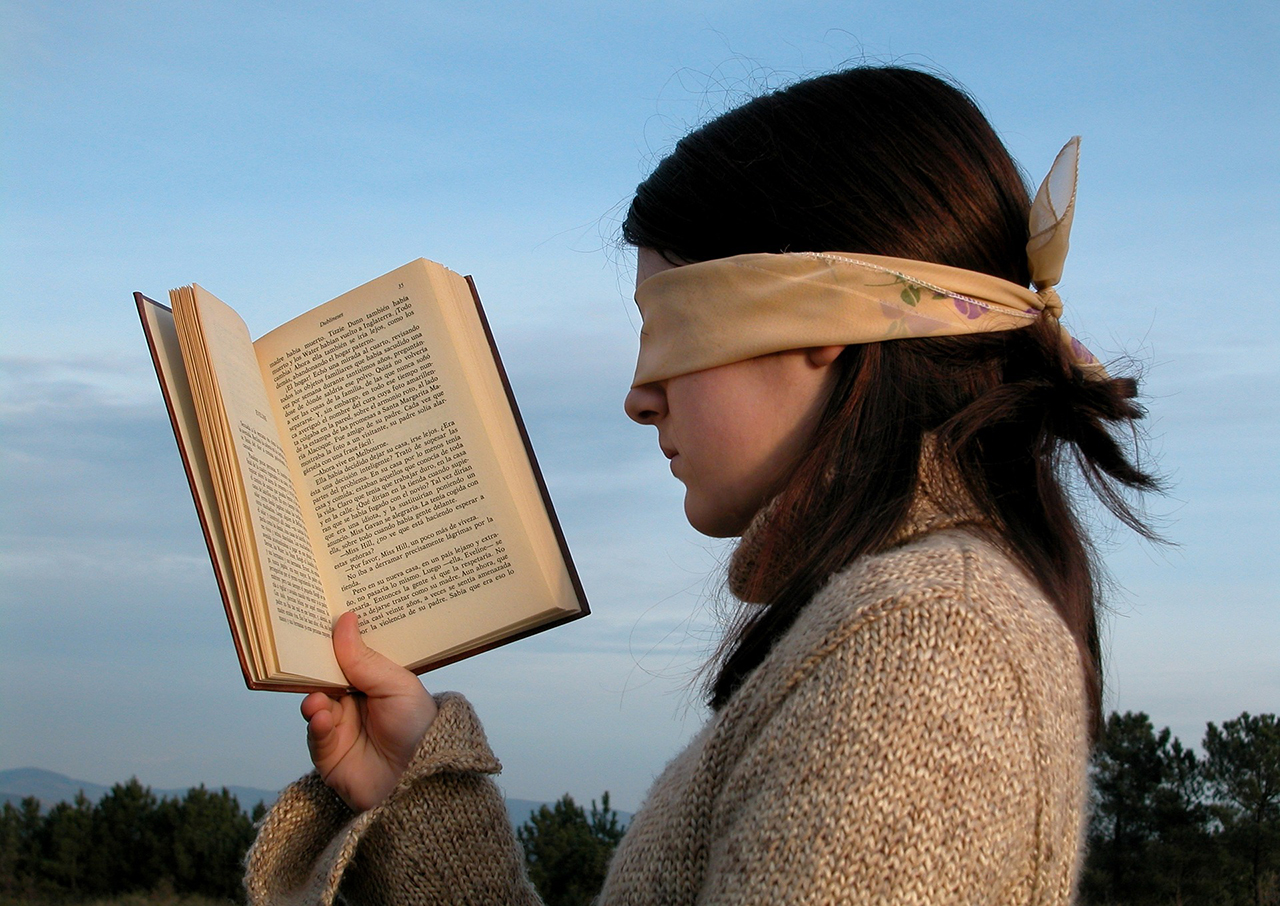 Uma menina em meio a natureza com um livro aberto na mão, mas seus olhos estão vendados por um pano