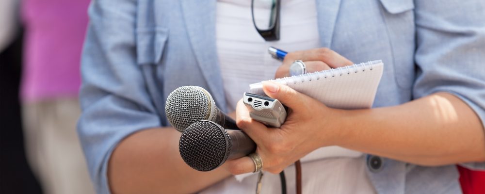 Jornalista com caderno de anotações, gravador de som e um microfone em mãos