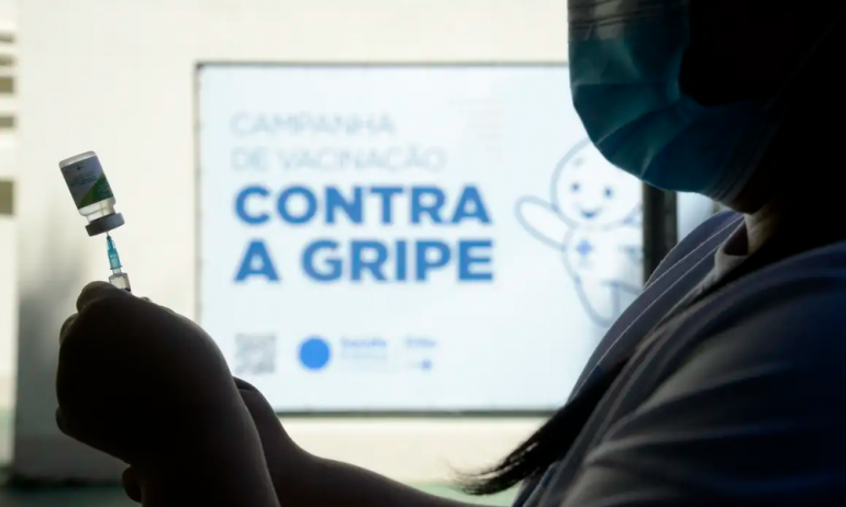 A foto mostra a silhueta de uma profissional da saúde segurando uma seringa com a vacina contra a gripe. Ao fundo está um cartaz om a imagem do personagem Zé Gotinha e a frase "CAMPANHA DE VACINAÇÃO CONTRA A GRIPE".