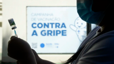 A foto mostra a silhueta de uma profissional da saúde segurando uma seringa com a vacina contra a gripe. Ao fundo está um cartaz om a imagem do personagem Zé Gotinha e a frase "CAMPANHA DE VACINAÇÃO CONTRA A GRIPE".
