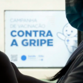 A foto mostra a silhueta de uma profissional da saúde segurando uma seringa com a vacina contra a gripe. Ao fundo está um cartaz om a imagem do personagem Zé Gotinha e a frase "CAMPANHA DE VACINAÇÃO CONTRA A GRIPE".